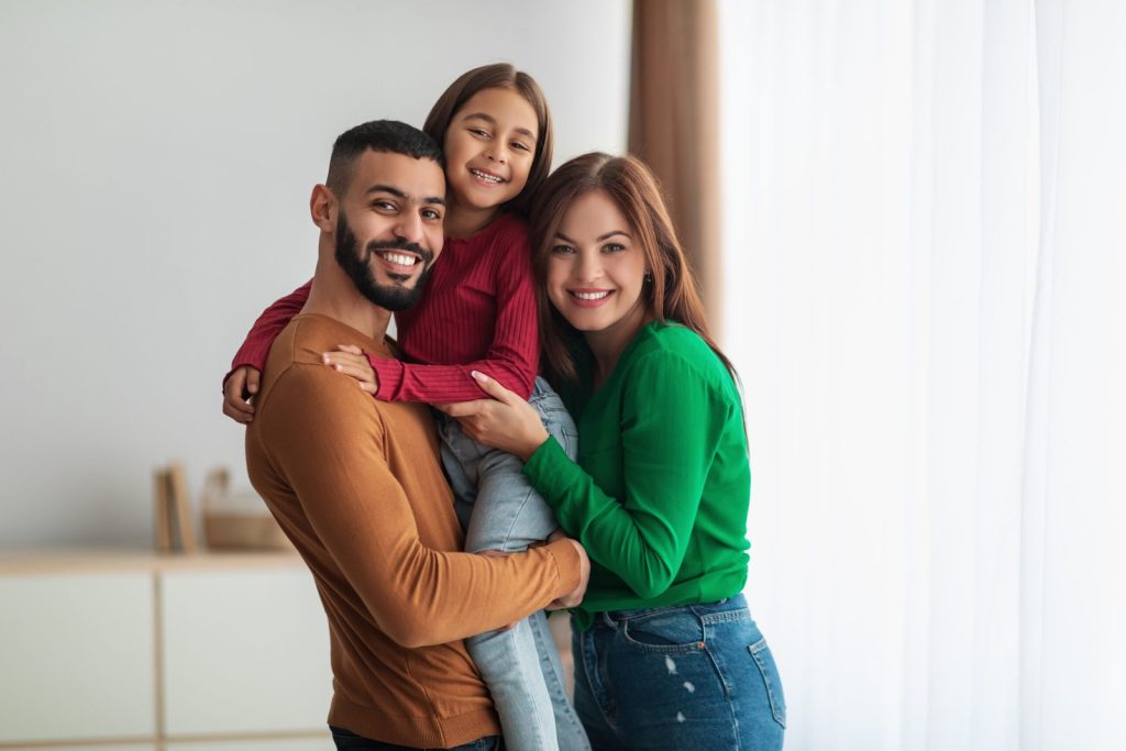 portrait-cheerful-arabic-family-three-people-hugging-standing-living-room-posing-photo-looking-camera-smiling-young-bearded-man-carrying-holding-his-daughter-hands-scaled